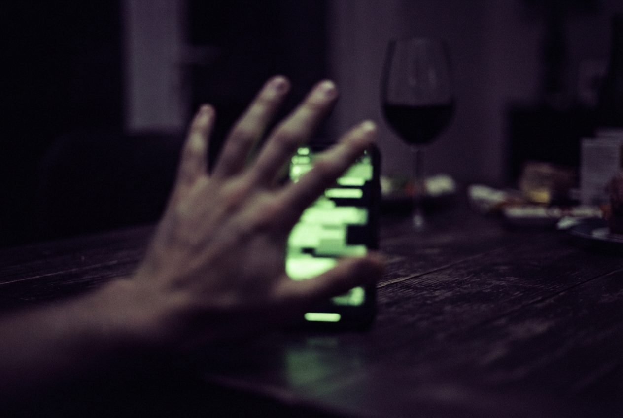 A blurred hand reaches toward a glowing green smartphone on a dark table beside a glass of red wine.